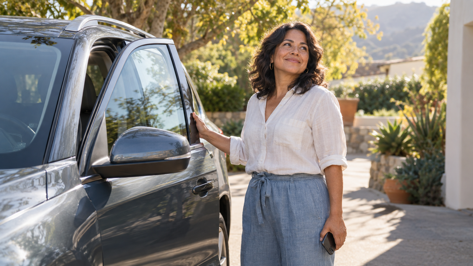 A smiling woman stands beside her gray SUV in a sunny driveway, one hand resting on the door.