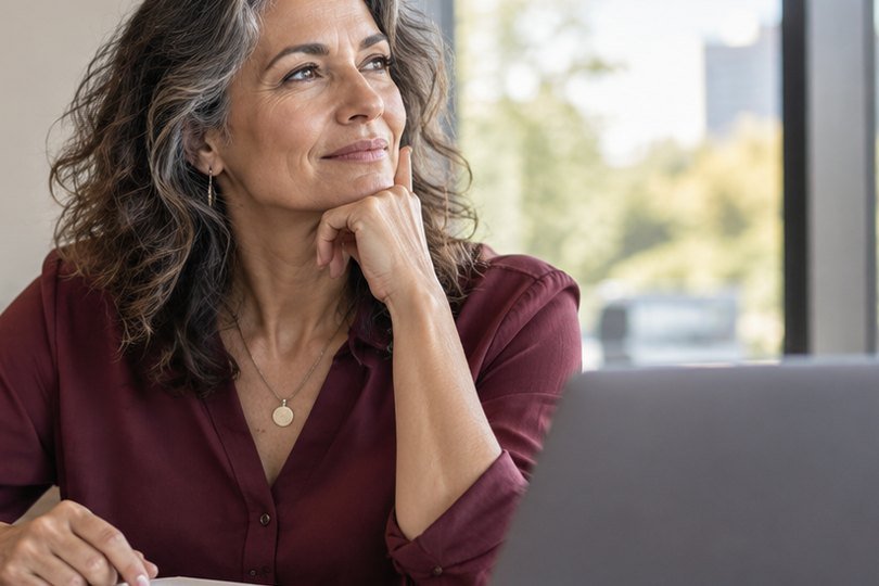 A woman in her late 40s pauses thoughtfully at her desk during the workday.